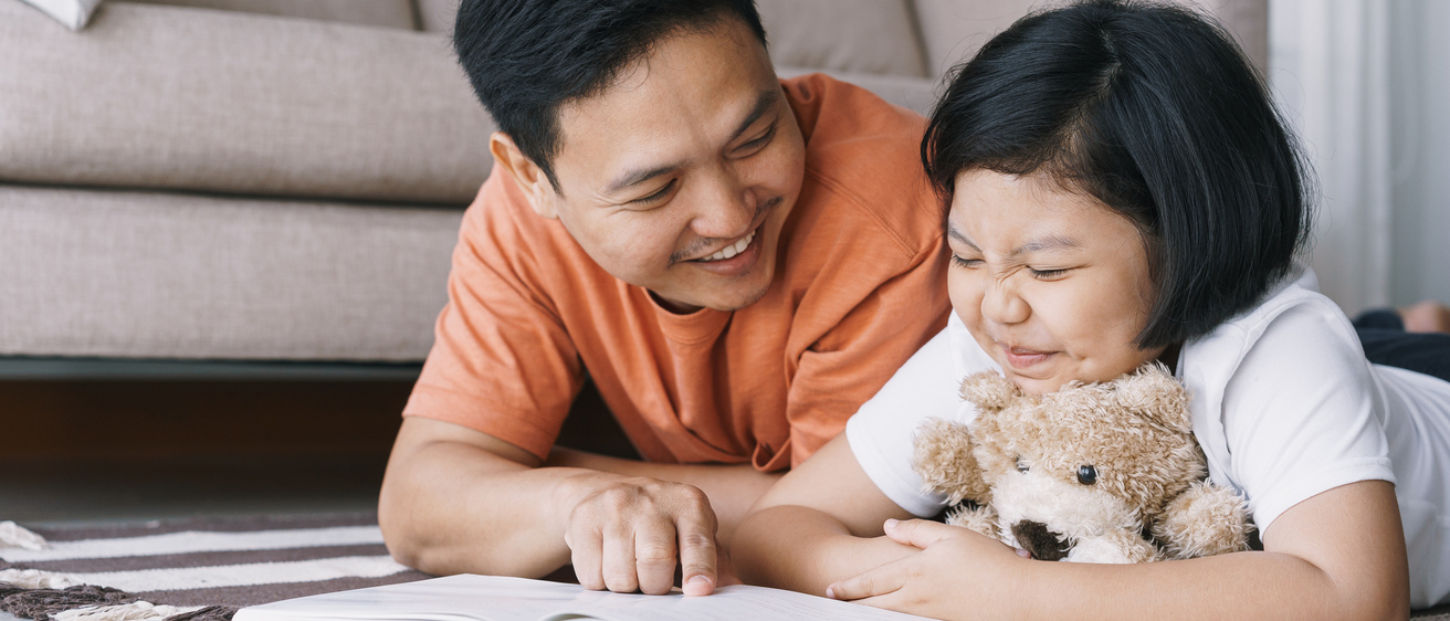 Asian father and daughter are reading book while lying on the carpet in the living room at her home. The cute little girl hugs the doll and smile happily with his father